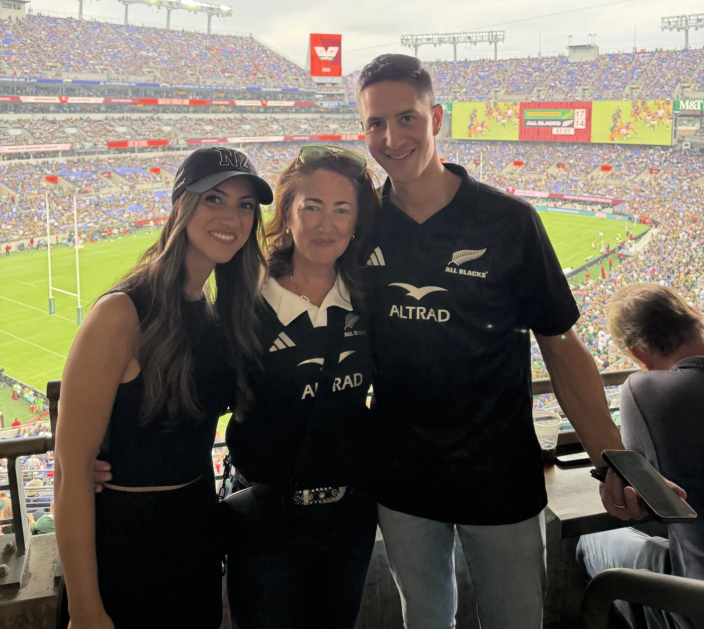 Three spectators posing together in the stands, with the rugby match behind them